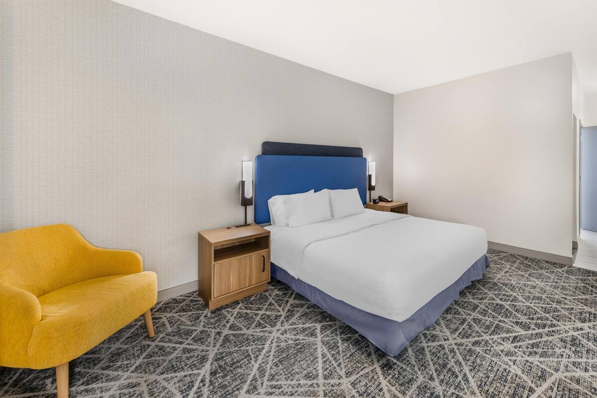 A hotel room with a bed, a nightstand, a wooden nightstand, a yellow armchair, and a patterned carpet.