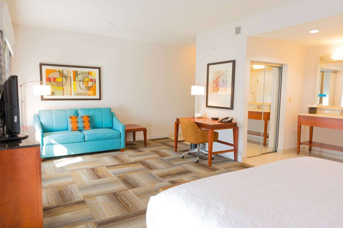A hotel room with a bed, a blue sofa with orange pillows, a wooden desk, a chair, a desk lamp, a telephone, and a framed picture on the wall.