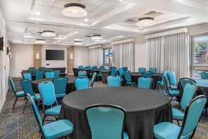 A conference room with round tables, blue chairs, a large screen, and a view of the outdoors. - Hampton Inn & Suites Riverside/Corona East (Riverside)