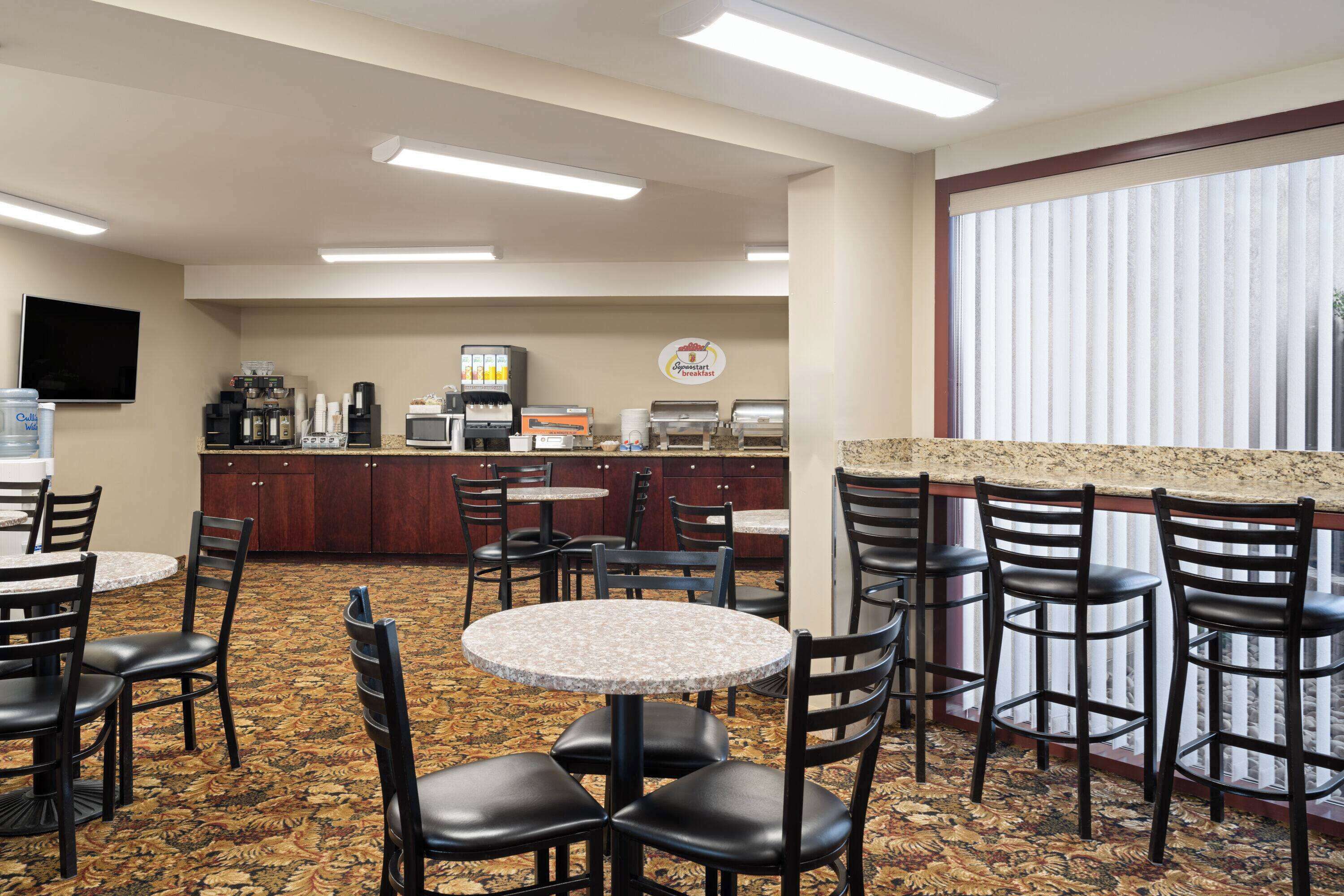 A hotel dining area with round tables and chairs, a kitchen in the background, and a large window with blinds.