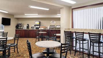 A hotel dining area with round tables and chairs, a kitchen in the background, and a large window with blinds.
