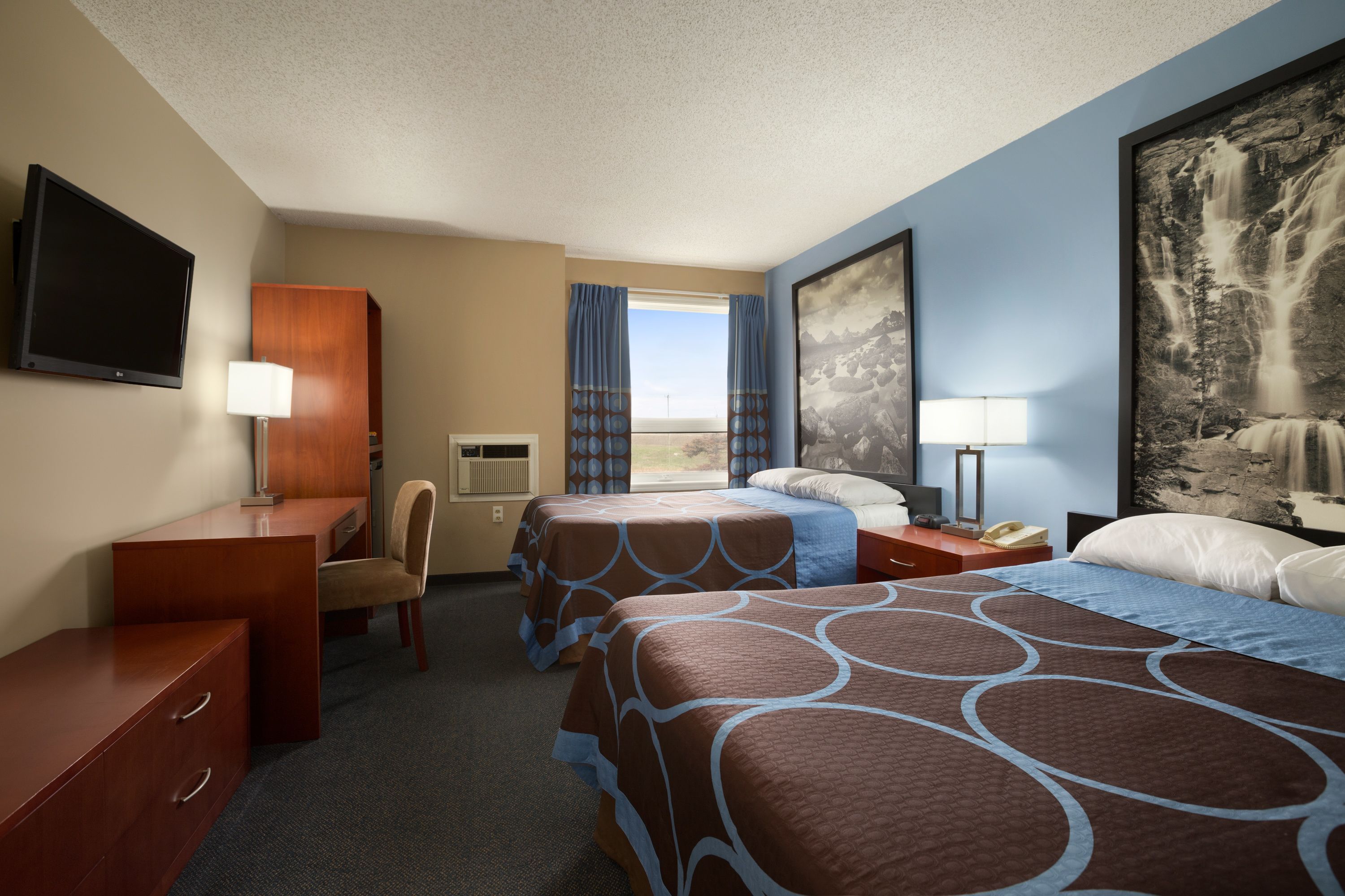 A hotel room with two beds, a desk, a chair, a TV, and a large window with curtains.