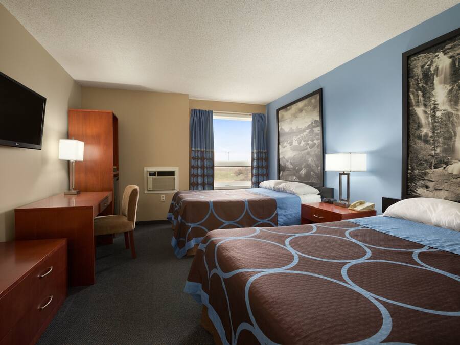 Quarto de hotel com duas camas, uma escrivaninha, uma cadeira, televisão e uma janela grande com cortinas.