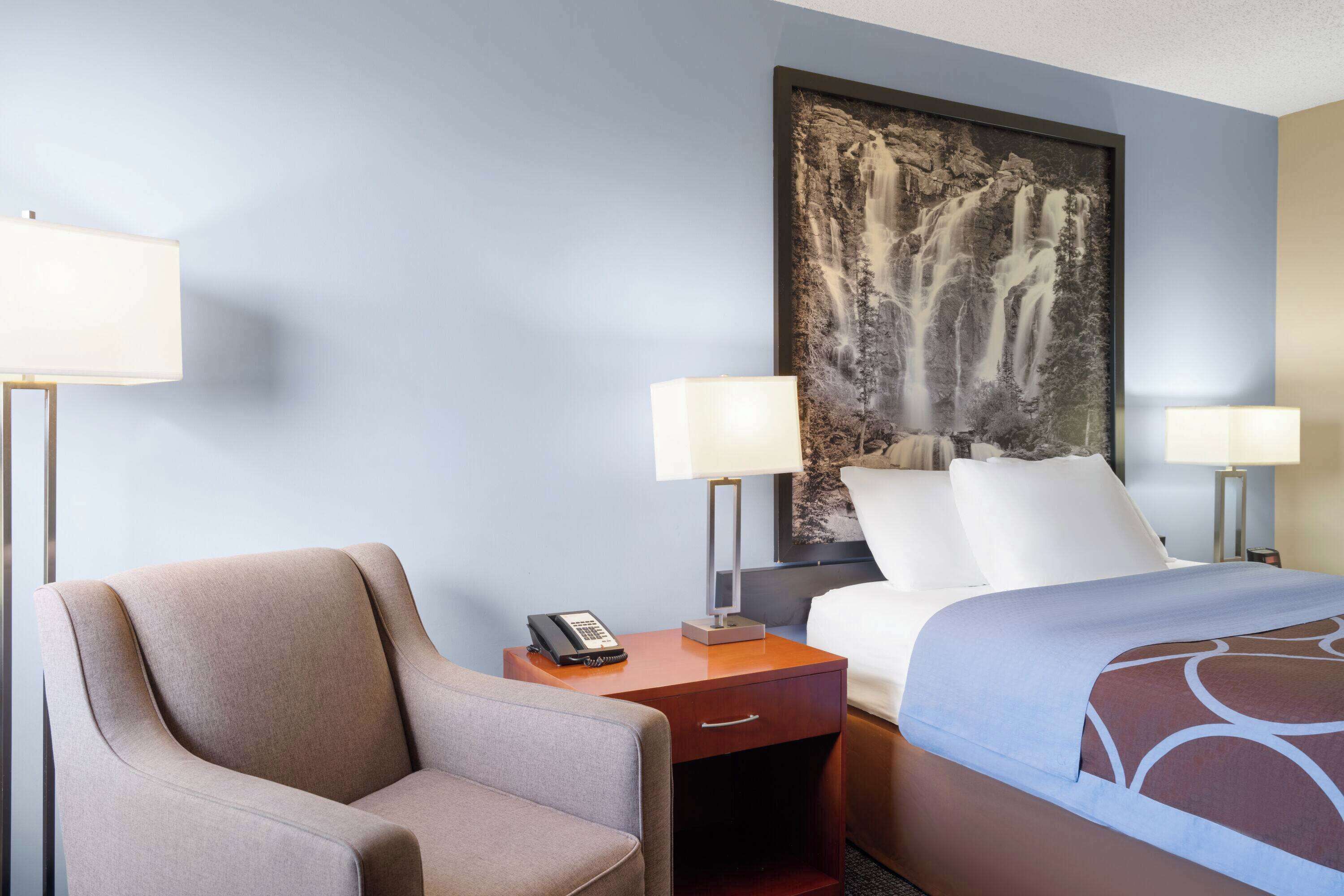 A hotel room with a bed, a chair, a nightstand with a phone, and a painting of a waterfall on the wall.