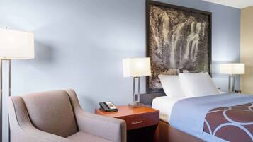 A hotel room with a bed, a chair, a nightstand with a phone, and a painting of a waterfall on the wall.