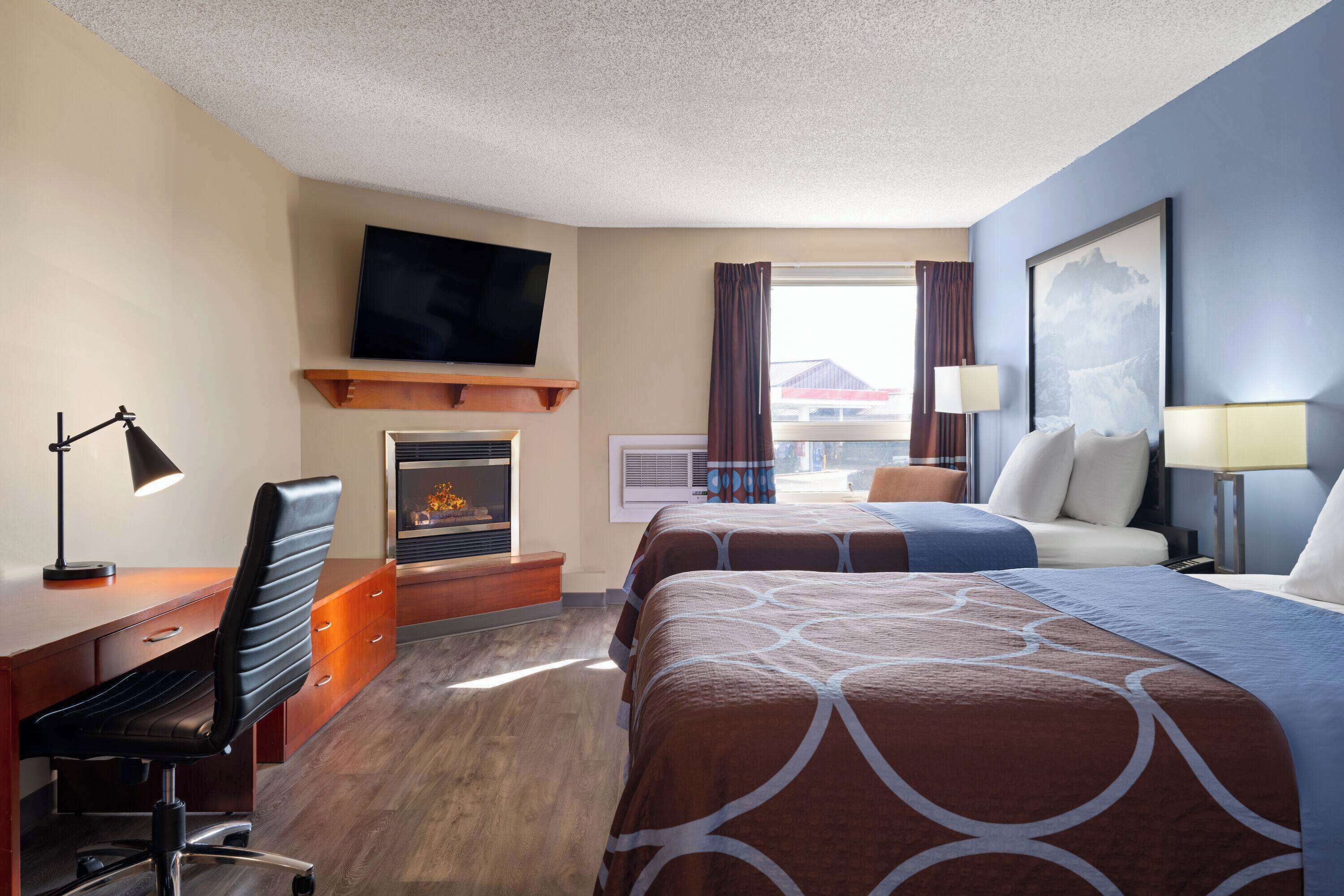 A hotel room with a bed, a desk with a chair, a TV, a fireplace, and a window with curtains.