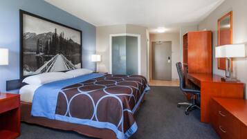 A hotel room with a large bed, a desk, a chair, a wardrobe, and a painting of a mountain landscape.