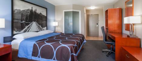 A hotel room with a large bed, a desk, a chair, a wardrobe, and a painting of a mountain landscape.
