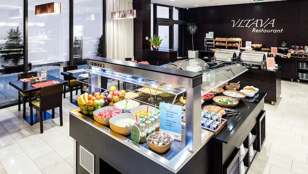 A buffet area with various food options, a sign indicating fruit and juice availability, and a modern interior design with a black wall and large windows. - Occidental Praha Five (Prague)