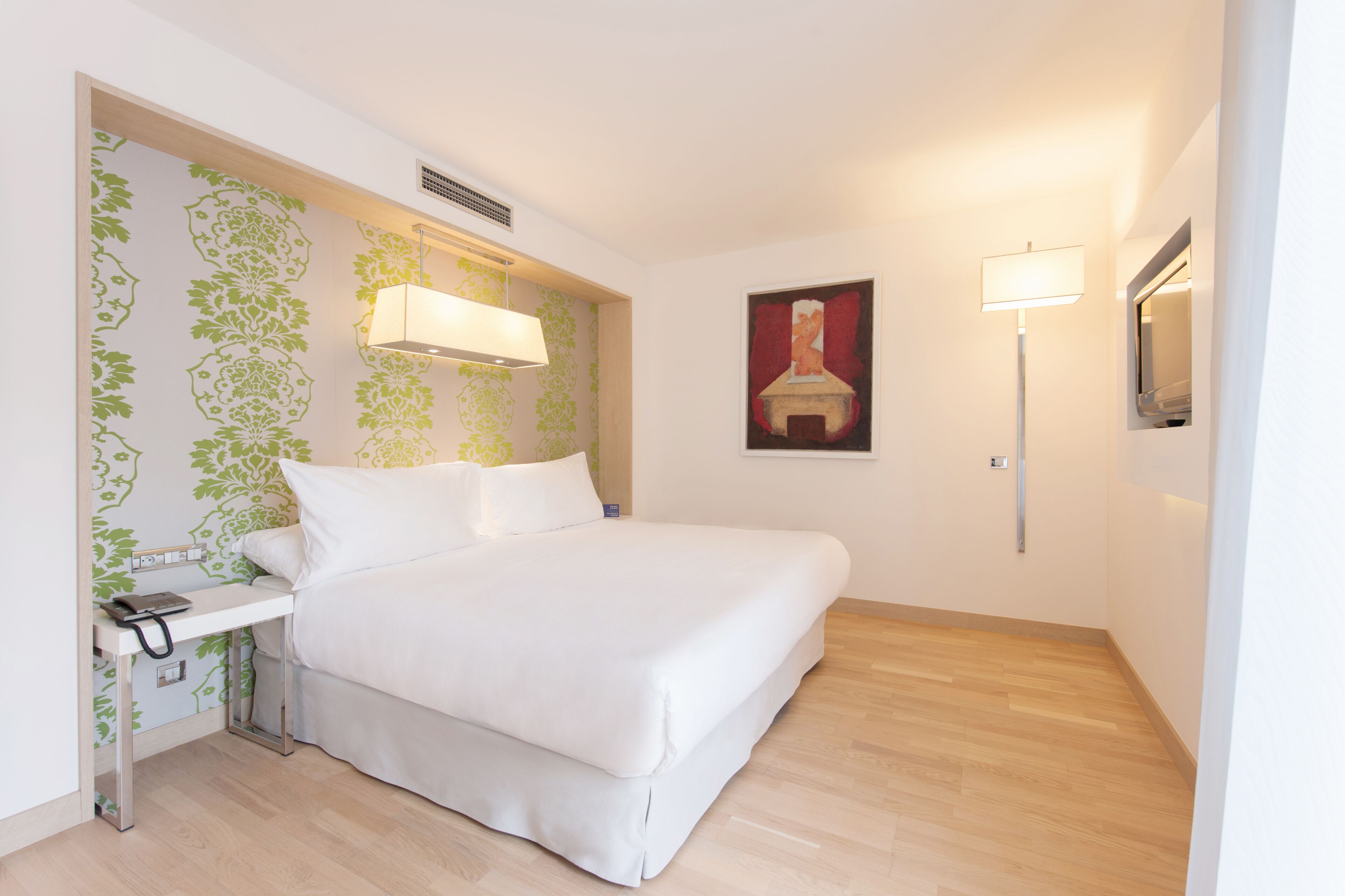 A hotel room with a bed, bedside table, lamp, and a painting on the wall.