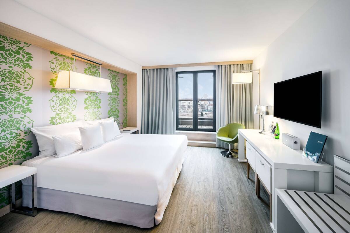 Habitación de hotel con una cama grande, un escritorio, una silla, un televisor y una ventana con cortinas.