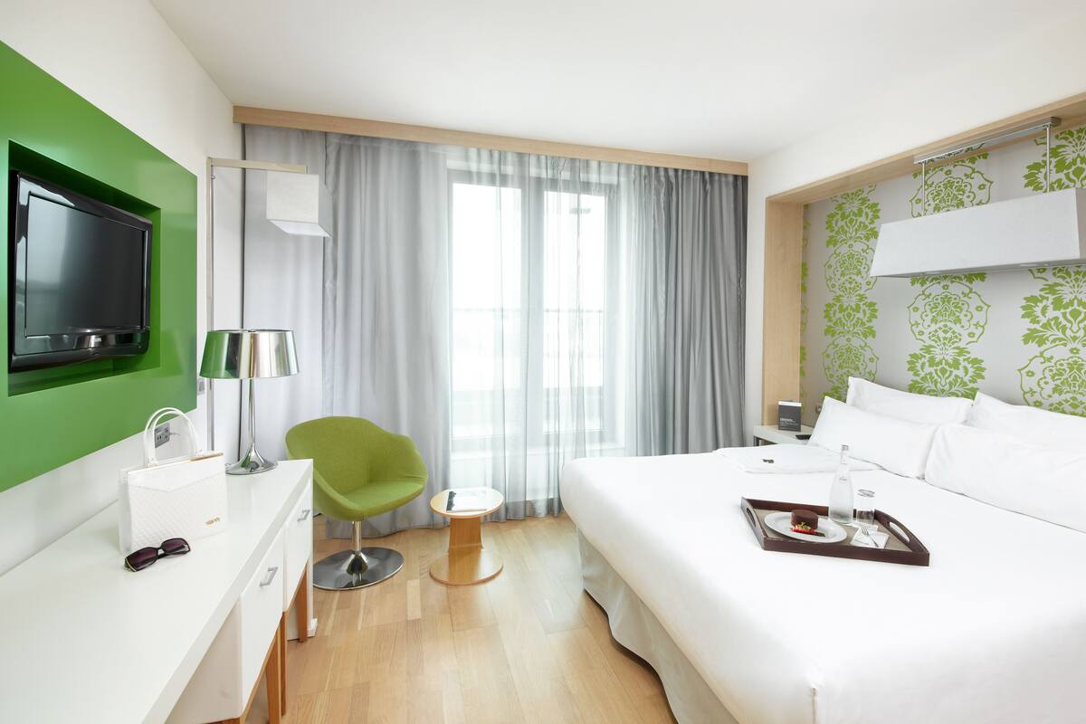 Una habitación de hotel con cama, escritorio, silla, televisión y ventana con cortinas.