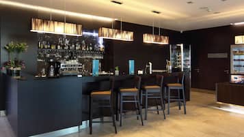Zona de bar moderna con mostrador oscuro, tres taburetes, lámparas colgantes y una barra bien surtida con varias botellas y vasos.