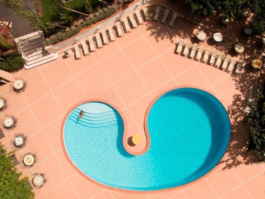 En bild tagen från luften som visar en pool med en ovanlig form, omgiven av solstolar och en kakelbeklädd terrass.