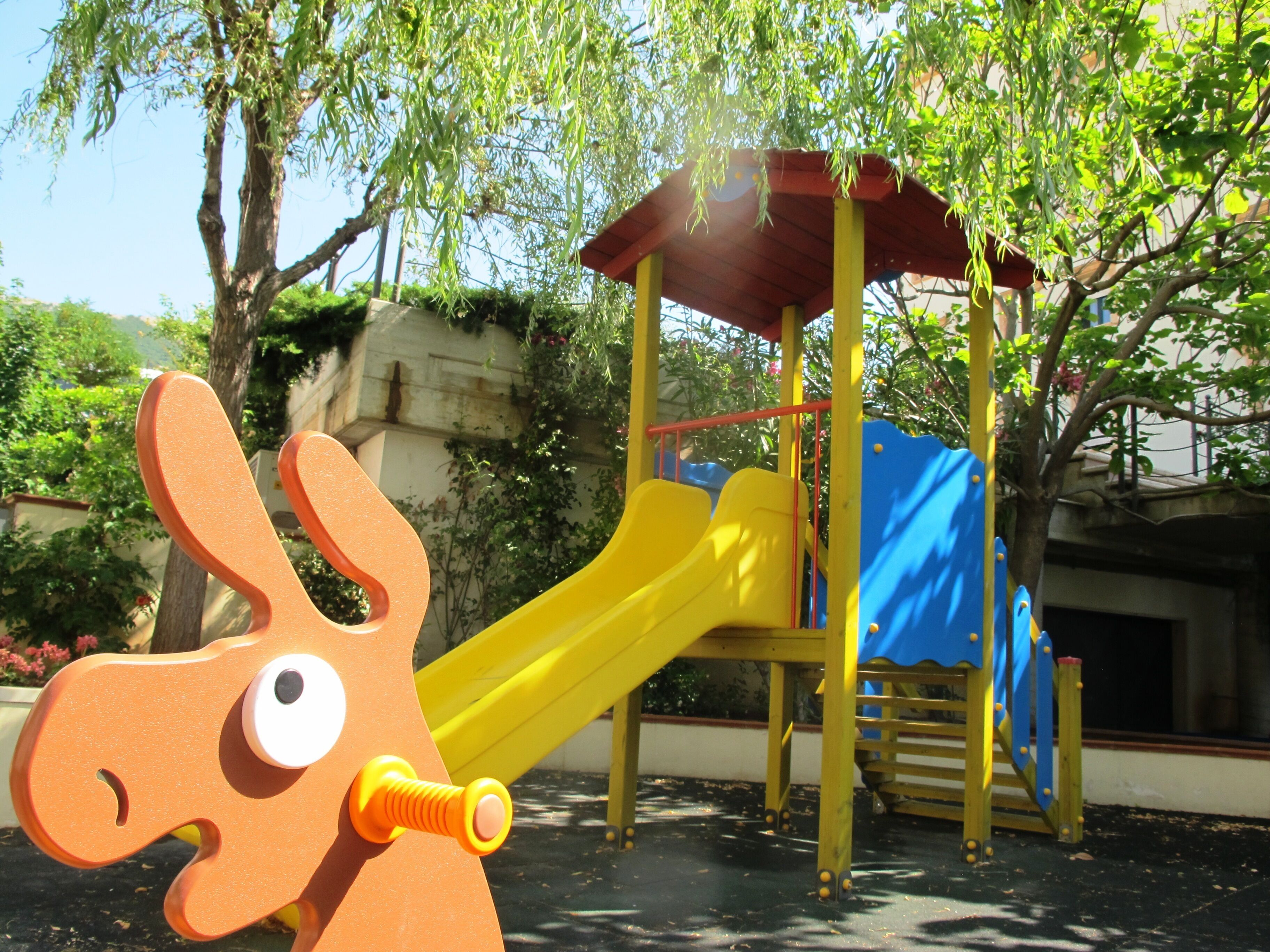 A playground with a slide and climbing structure, surrounded by trees and a stone wall.