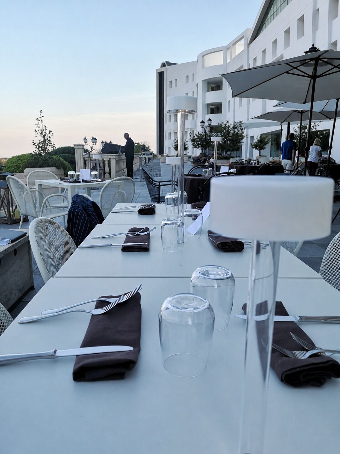 An outdoor dining area with tables set for two, chairs, and umbrellas.