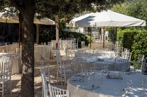 Un área de comedor al aire libre con mesas redondas y sillas blancas, rodeada de árboles y cubierta por grandes sombrillas blancas.