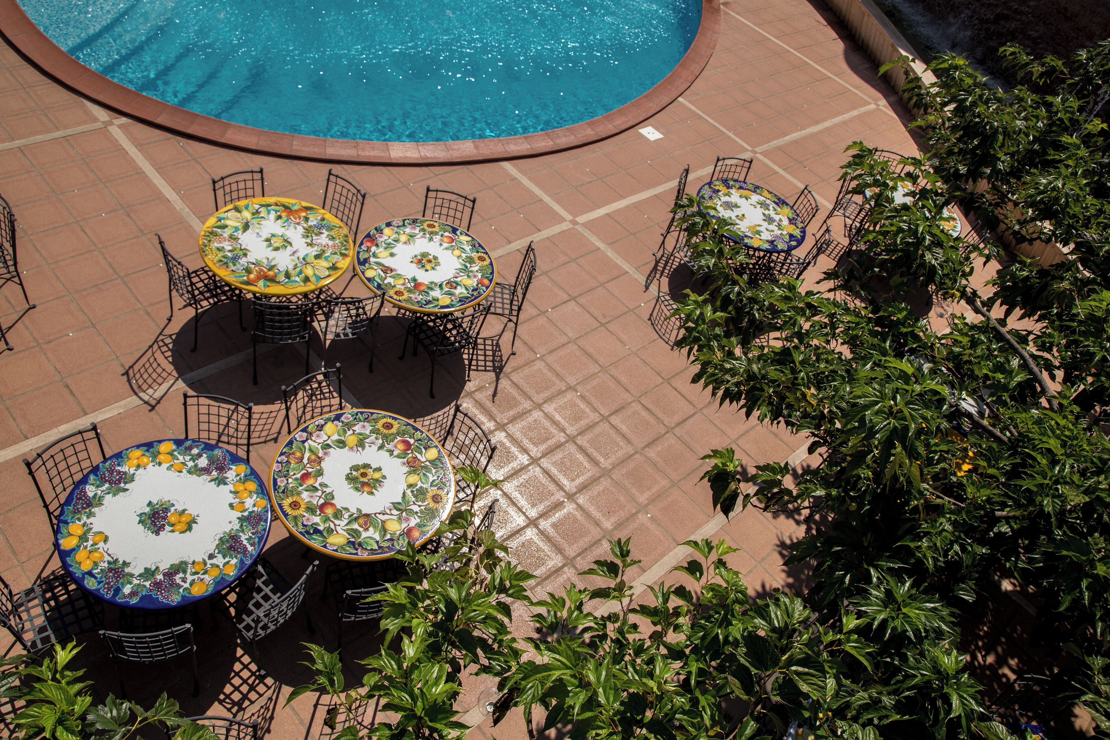 An outdoor pool area with multiple round tables and chairs, surrounded by greenery.
