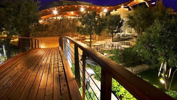 Una terraza de madera con mobiliario exterior, rodeada de vegetación y un edificio con arquitectura iluminada.