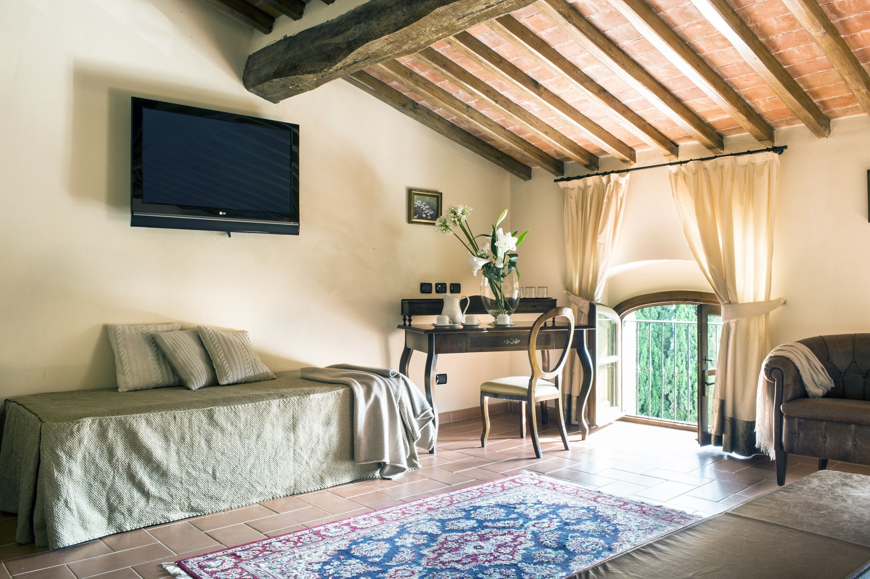 Una habitación acogedora con un sofá, un escritorio con silla, un televisor montado en la pared y vistas a través de una ventana con cortinas.