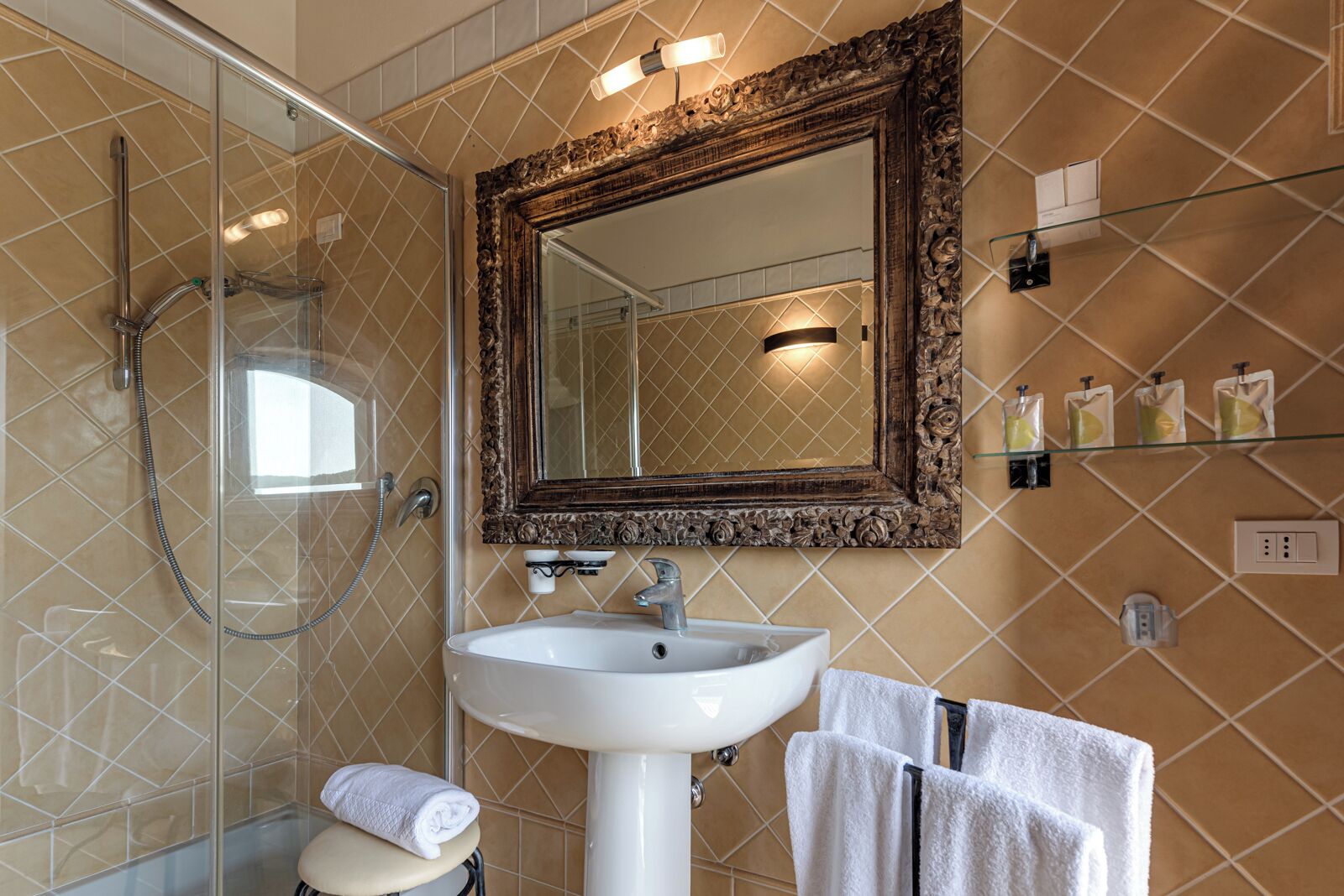 A bathroom with a glass-enclosed shower, a white sink, a large ornate mirror, and a shelf with toiletries.