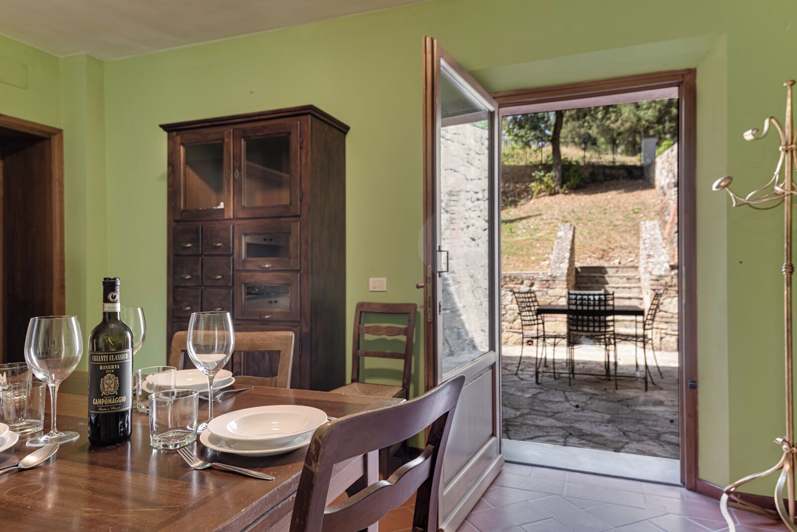 Un comedor con una mesa preparada para dos personas, una botella de vino y vistas a un patio exterior con una mesa y sillas.