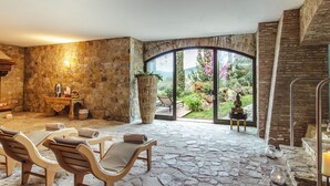 A spa area with stone flooring, wooden chairs, a table with amenities, and a view of a garden through a glass door. - Hotel Villa Campomaggio (Radda in Chianti)