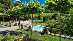 A landscaped garden with a swimming pool, a vineyard in the background, and a stone pathway. - Hotel Villa Campomaggio (Radda in Chianti)