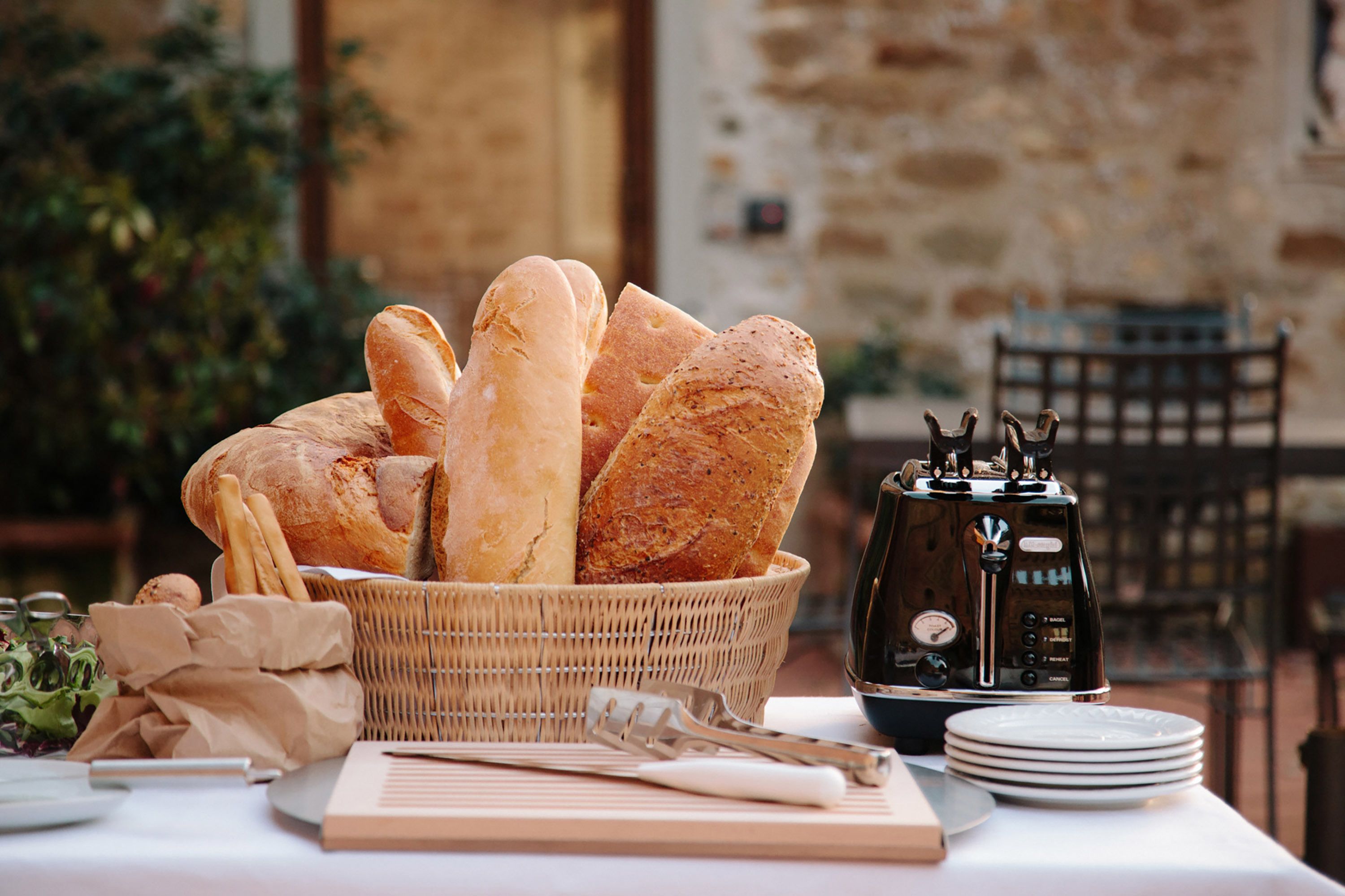 Un panier en osier rempli de baguettes est posé sur une table, à côté d’une machine à café et d’assiettes.