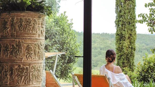 A stone pillar with carvings, a glass door leading outside, and a person sitting on a lounge chair. - Hotel Villa Campomaggio (Radda in Chianti)