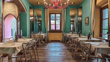 Een eetzaal met meerdere tafels en stoelen, een kroonluchter en grote ramen.