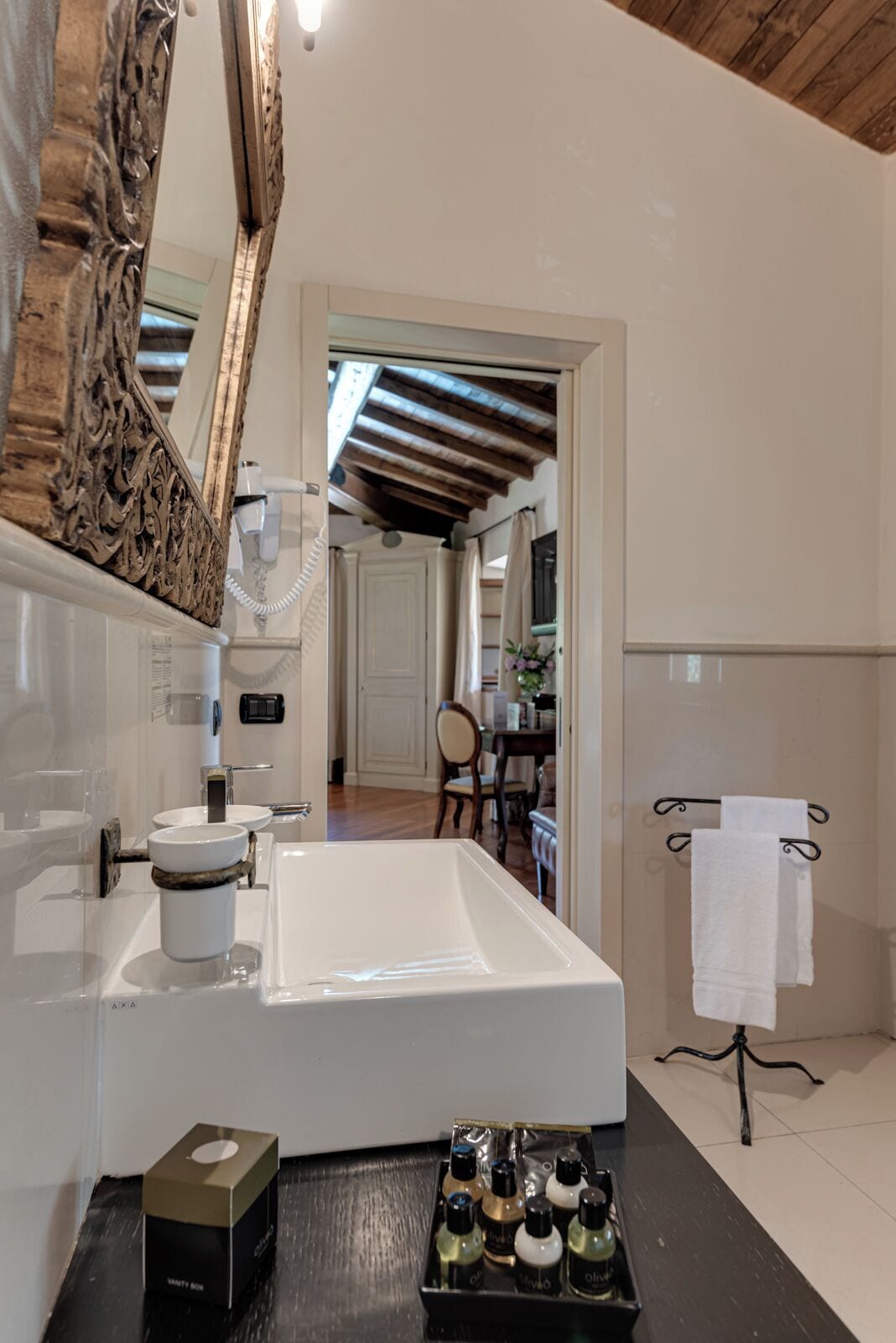 Een badkamer met een groot wit wastafel, een spiegel met een sierlijk frame en een zwart aanrechtblad met een doos toiletartikelen.