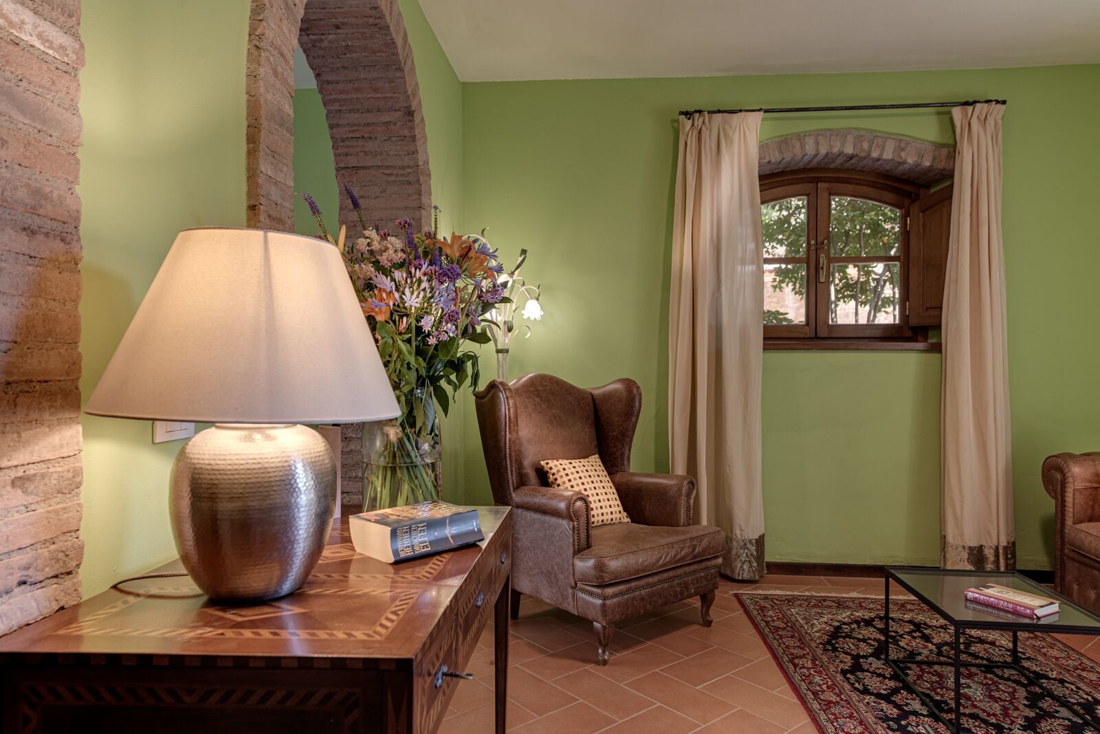 A living room with a leather armchair, a table with a lamp, a vase with flowers, and a window with curtains.