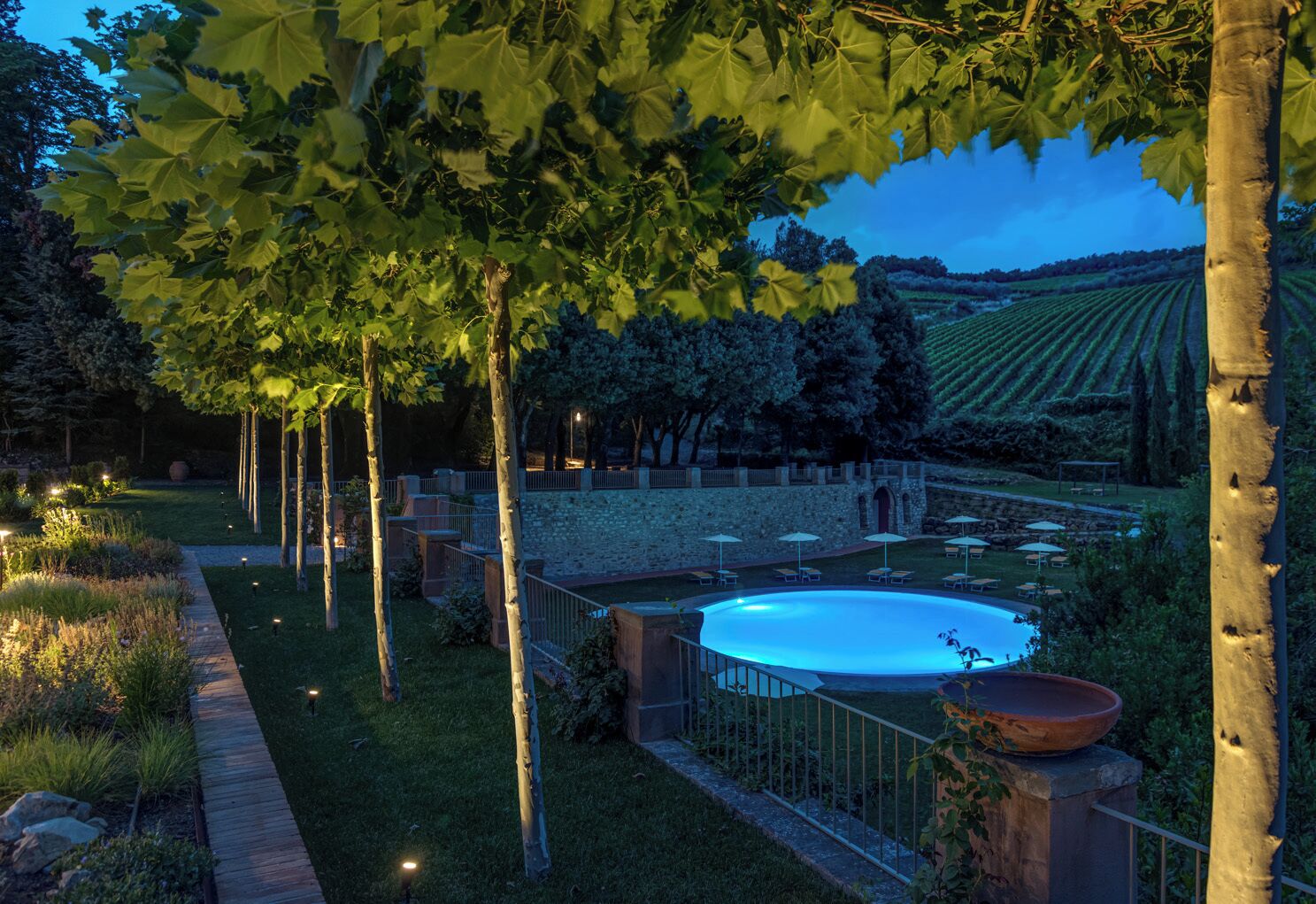 A pool surrounded by a garden with trees and a view of vineyards.