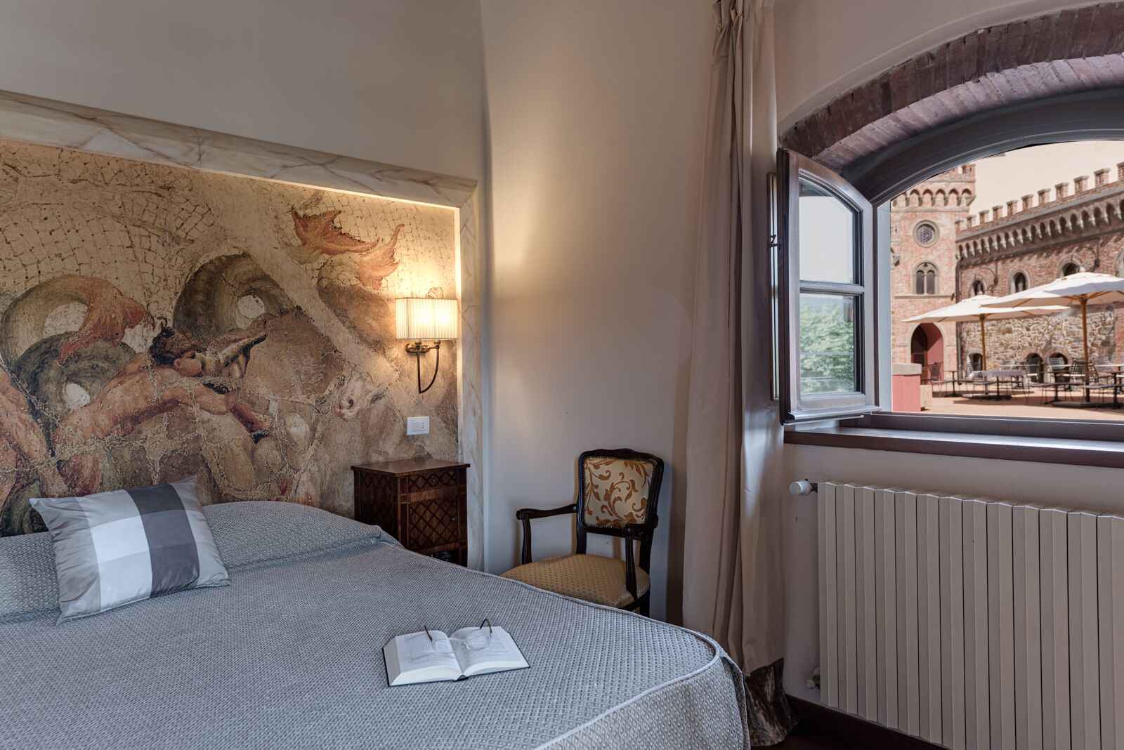 A hotel room with a bed, a reading lamp, a chair, and a view of an outdoor courtyard with stone architecture.