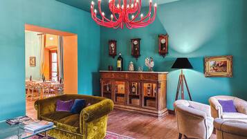 A room with teal walls, a wooden cabinet with bottles, a green sofa, a white armchair, and a red chandelier.