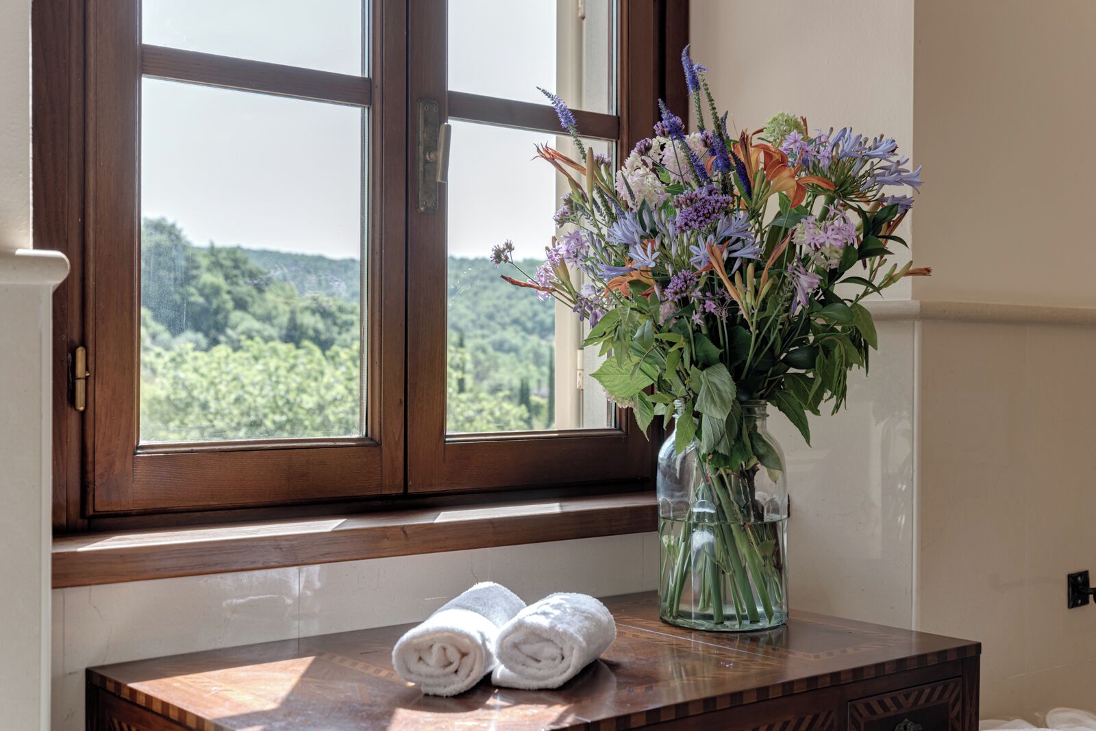 Una ventana con vistas al verdor, un florero con flores y toallas enrolladas sobre una superficie de madera.