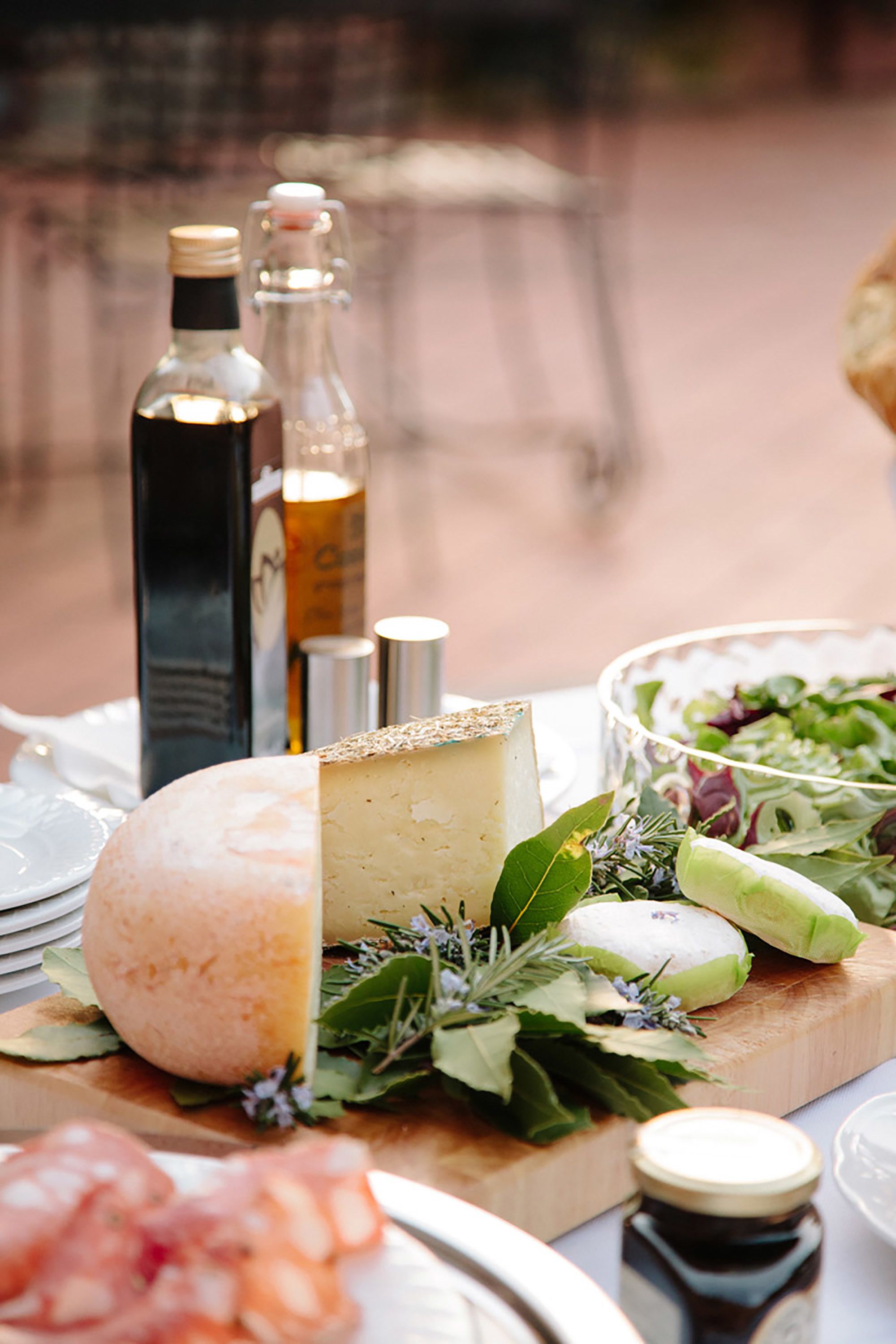 Una tabla de quesos con diferentes tipos de queso, una botella de aceite de oliva y un bol de ensalada sobre una tabla de madera.