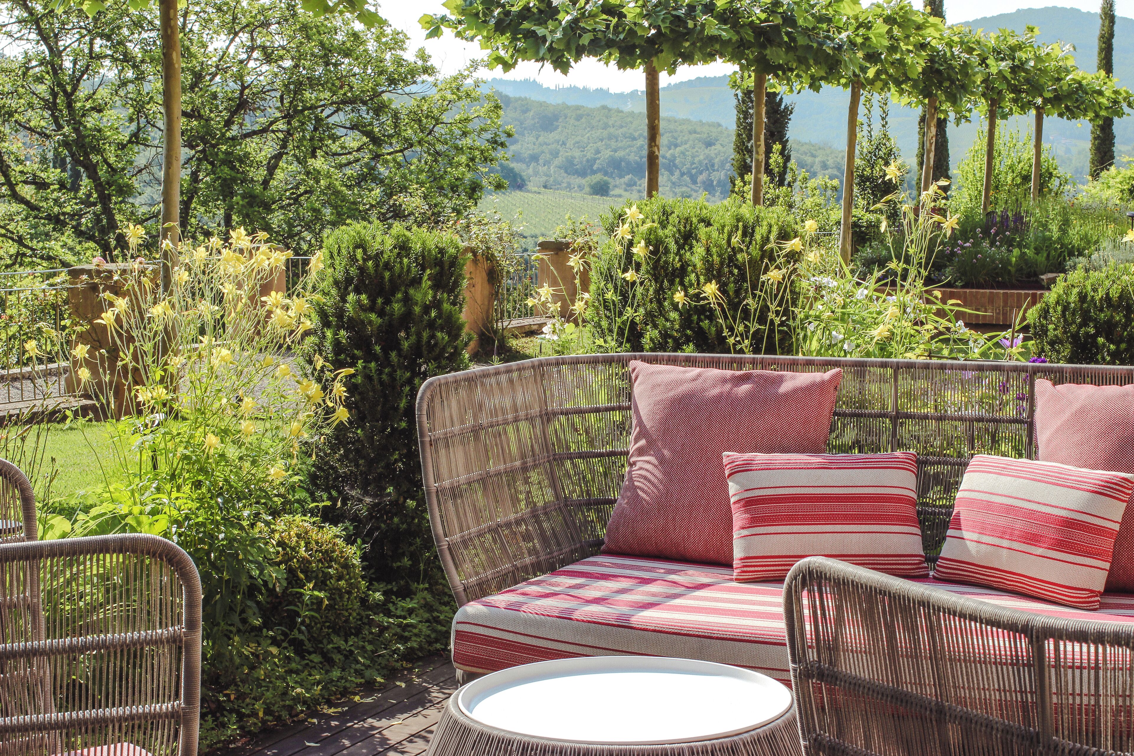 Un canapé en osier agrémenté de coussins à rayures rouges et blanches, une table ronde et un jardin luxuriant où poussent différentes espèces de plantes, le tout offrant une vue sur des collines lointaines.