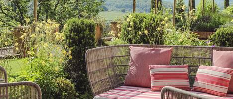 Un canapé en osier agrémenté de coussins à rayures rouges et blanches, une table ronde et un jardin luxuriant où poussent différentes espèces de plantes, le tout offrant une vue sur des collines lointaines.