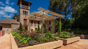 Un patio con un edificio de piedra, una pérgola de madera y bancales elevados con diversas plantas.