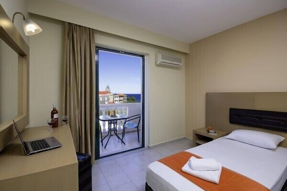 A hotel room with a bed, a desk with a laptop, a bottle of wine, and a table with chairs on a balcony.