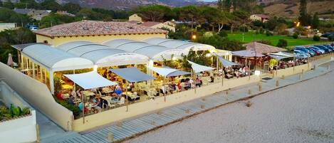 A coastal restaurant with outdoor seating, a covered walkway, and a view of the beach and mountains.