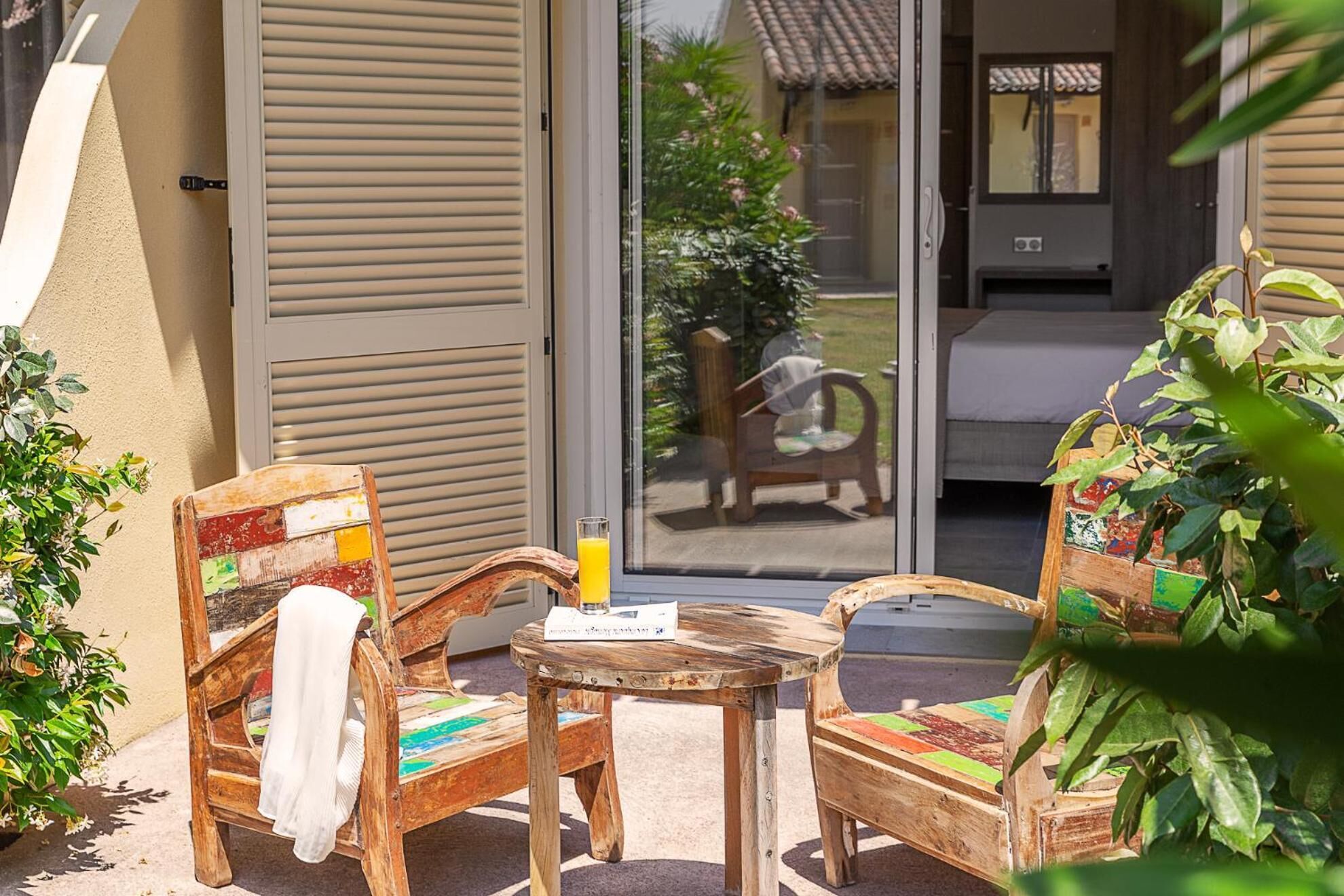 Un espace extérieur aménagé avec des meubles en bois, comprenant une table et deux chaises, un verre de jus d’orange et une vue sur une maison, visible à travers une porte coulissante.