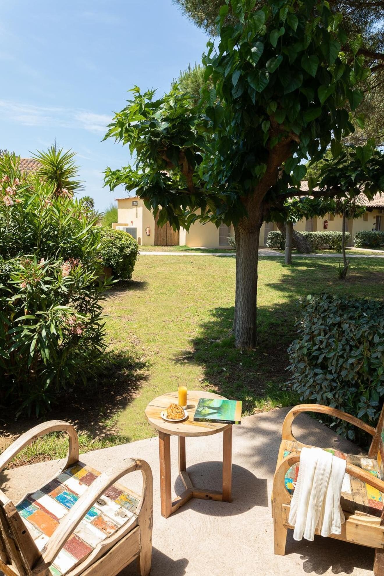 Un patio con due sedie di legno, un tavolino rotondo con un libro e un bicchiere di succo d'arancia, e un albero che offre ombra.
