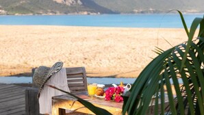 A wooden bench with a hat and a table set with a drink, flowers, and a book, overlooking a beach and mountains. - Hotel Marina di Lava (Appietto)