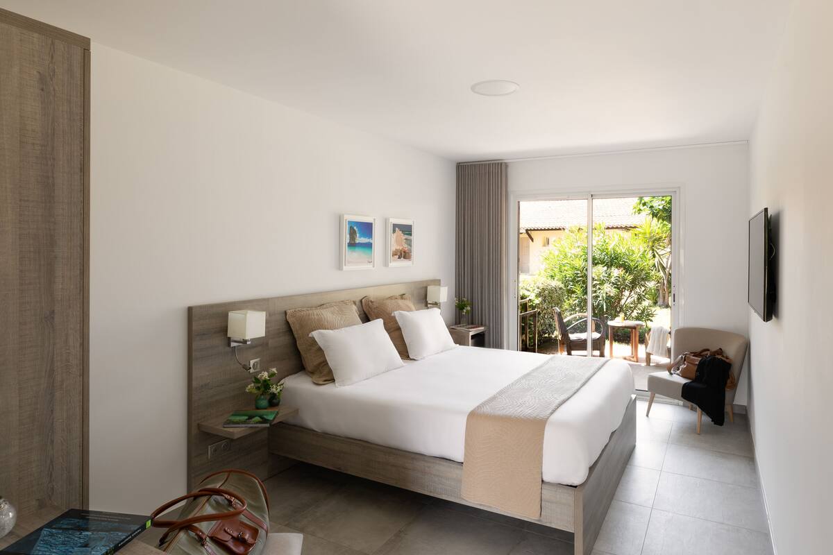 A modern hotel room with a large bed, a bedside table with a lamp, a TV mounted on the wall, and a view of the outdoors through a sliding door.