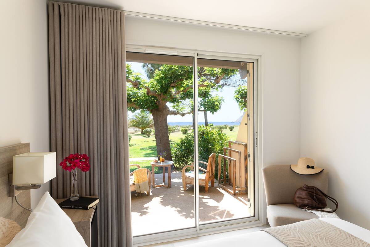 A hotel room with a large sliding glass door leading to an outdoor area with a view of the sea and greenery.