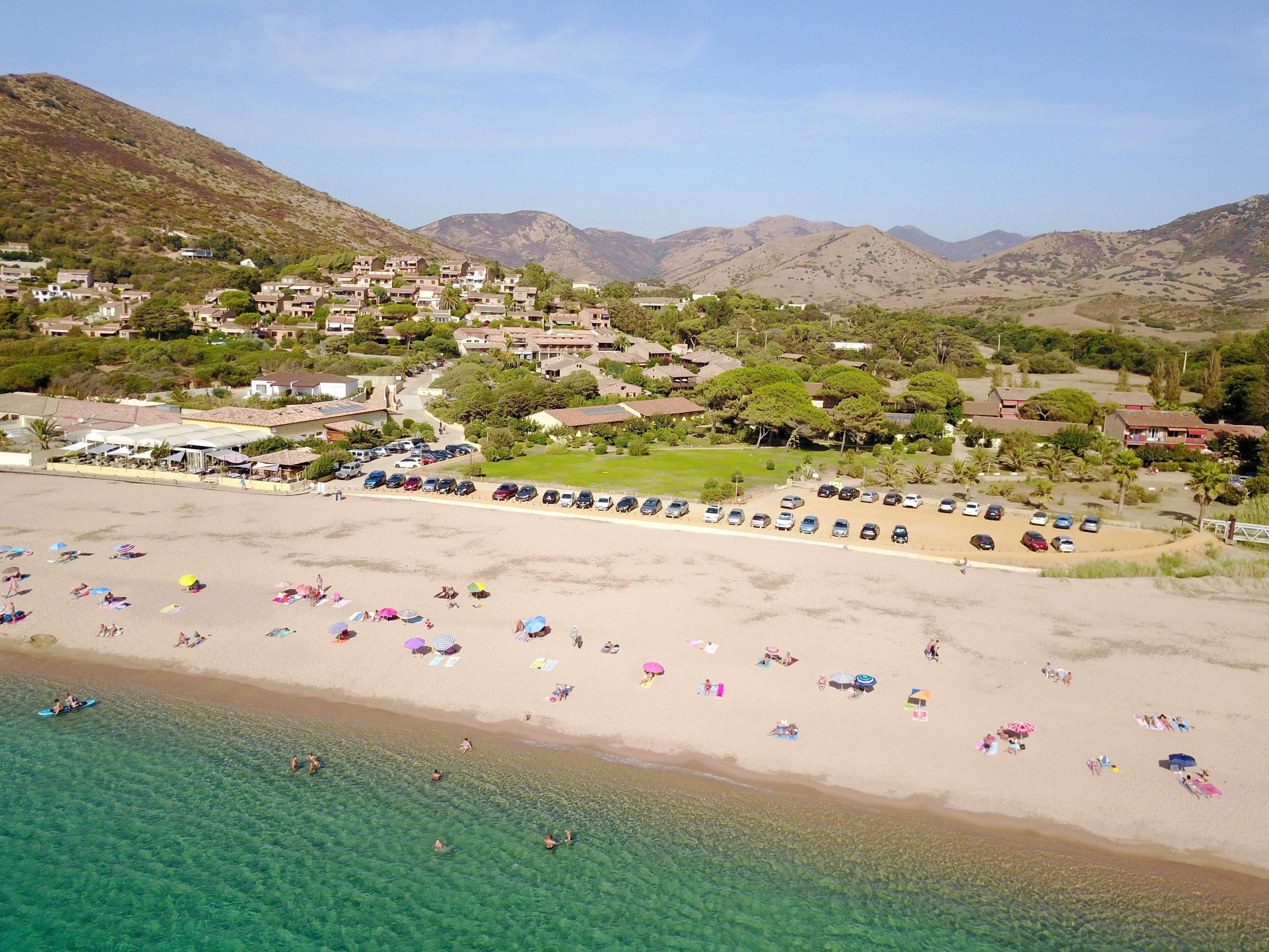 Una zona costera con playa, agua cristalina y un área residencial con casas y vegetación al fondo.
