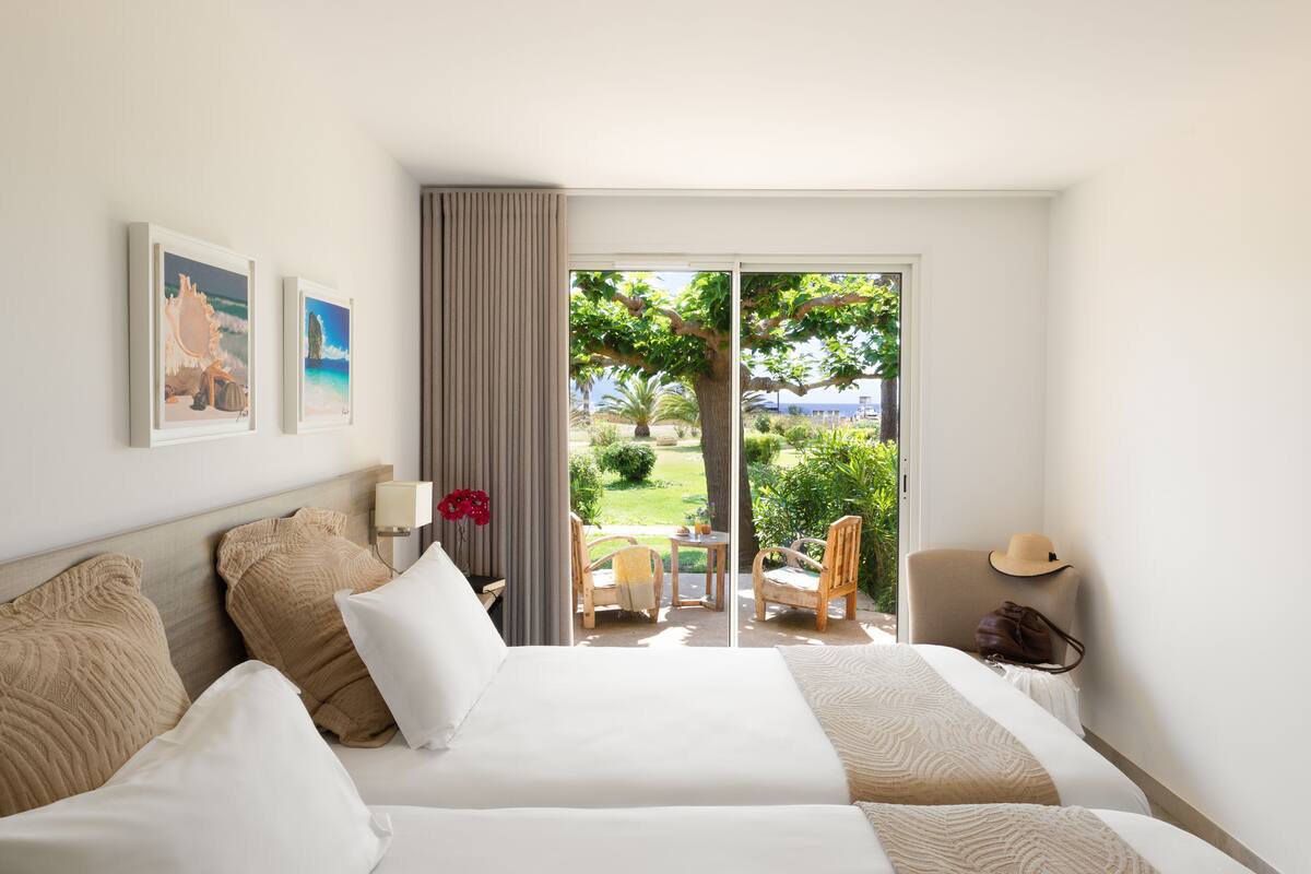 A hotel room with a large bed, a desk, and a view of a garden and sea through a sliding door.